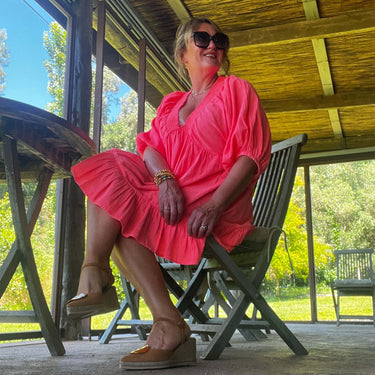 Woman in a bright pink dress sitting on a chair under a wooden canopy with trees in the background.