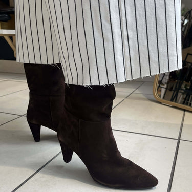 Dark brown suede ankle boots with pointed toes and low block heels, worn with a white and black striped skirt on a tiled floor.