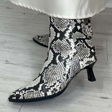 Snakeskin-patterned ankle boots worn with white pants on a light wooden floor.