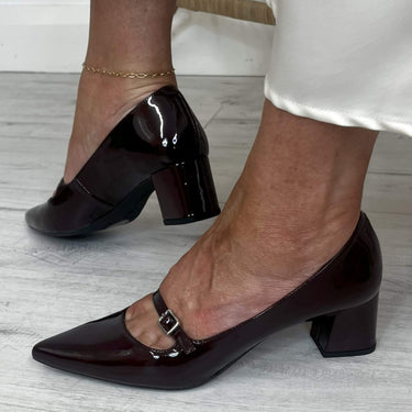 Black patent leather shoes with block heels worn by a person on a light wooden floor.