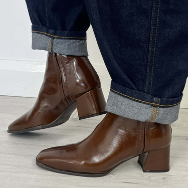 A person wearing dark brown pointed-toe ankle boots with block heels, paired with dark blue cuffed jeans, standing on a light gray wood floor.
