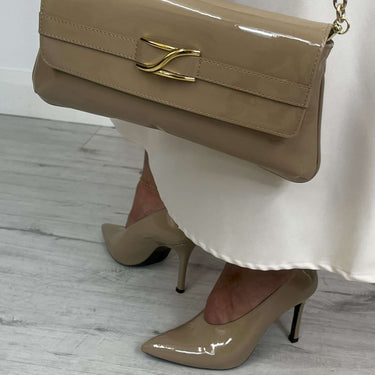 Beige handbag with gold chain strap held by a person wearing beige high heels on a light wooden floor.