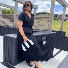 Woman in a black outfit holding a white bag with black stripes, standing outdoors.