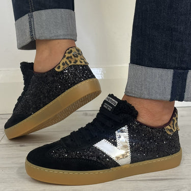 A person wearing black glitter sneakers with leopard print heel accents, silver side stripes, and gum soles, paired with dark cuffed jeans on a light wood floor.