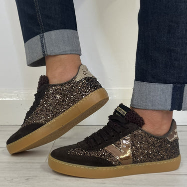 A person wearing dark brown and black glitter sneakers with a metallic rose gold side stripe and tan gum soles, paired with cuffed dark blue jeans on a light wood floor.