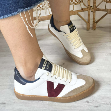 White sneakers with a maroon 'V' logo worn by a person on a light wooden floor.