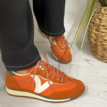 Orange sneakers worn with black pants on a light wooden floor.