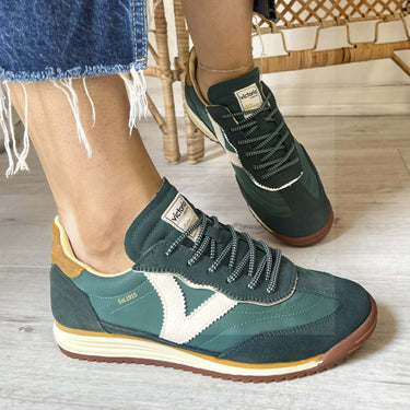Green and brown sneakers worn with denim shorts on a wooden floor.