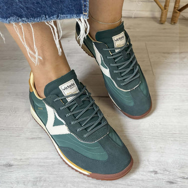 Green sneakers with brown soles worn by a person on a light wooden floor.
