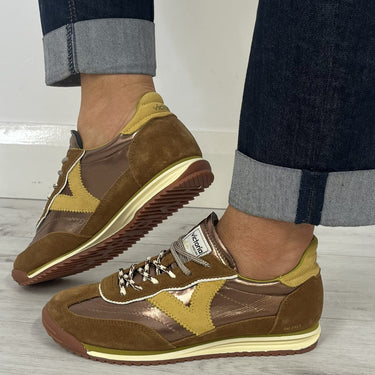 A person wearing brown and tan suede and leather sneakers with dark blue cuffed jeans, standing on a light gray wood floor.