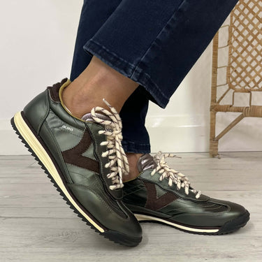 A person wearing dark olive green leather sneakers with brown side panels, cream laces, and a cream midsole, paired with dark blue jeans, standing on a light wood floor.