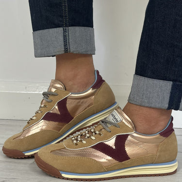 A person wearing cuffed dark blue jeans and metallic gold Vionic sneakers with burgundy accents, standing on a light gray wood floor against a white wall.