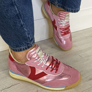 Pink and red sneakers worn with blue jeans on a light wooden floor.