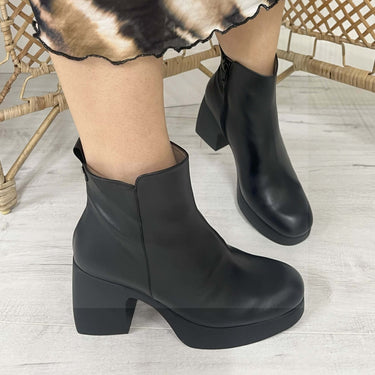 Black ankle boots worn by a person on a light wooden floor.