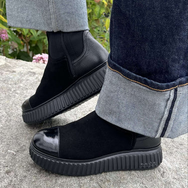 Black platform chunky boots with a glossy toe cap, worn with cuffed dark blue jeans on a stone surface with outdoor flowers in the background.