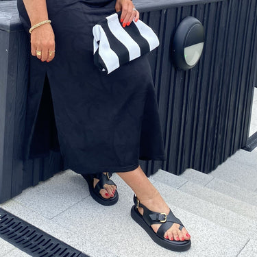 Woman in a black dress with sunglasses and a striped bag standing outdoors.