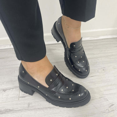 Black loafers with stud details worn with black pants on a light wooden floor.