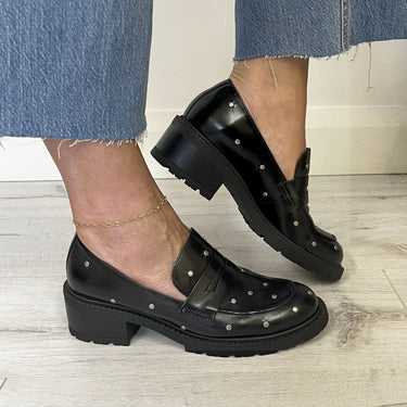 Black leather shoes with studded details worn by a person on a wooden floor.