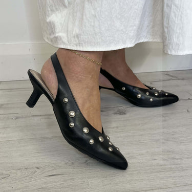 Black studded high-heeled shoes worn with a white skirt on a wooden floor.