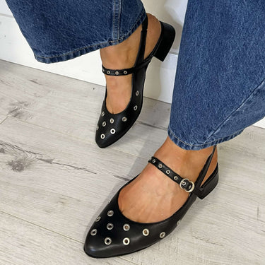 Black studded flats worn with blue jeans on a light wooden floor.