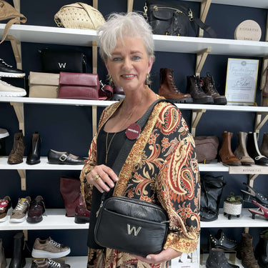 Woman holding a black handbag with a visible brand logo in front of shelves displaying various bags and shoes.
