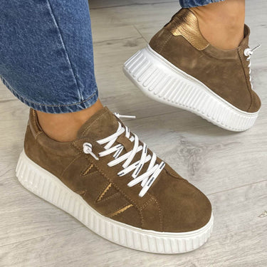 A pair of brown suede low-top sneakers with chunky white platform soles and white laces, shown being worn with blue jeans on a light gray wood floor.