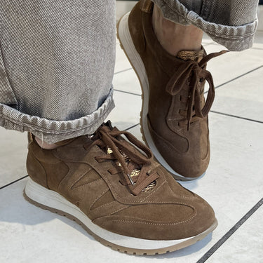 A person wearing brown suede sneakers with light gray cuffed jeans, standing on a light gray tiled floor.