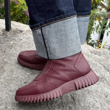 A person wearing dark blue jeans with the cuffs rolled up, standing on a large gray stone outdoors. The person is wearing a pair of low-top burgundy leather boots with chunky, textured soles.