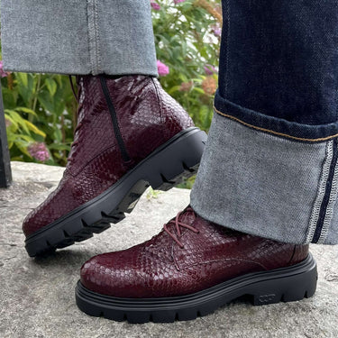 A close-up of a person wearing dark burgundy snakeskin-textured lace-up boots with black lug soles, paired with cuffed blue jeans, standing on a stone surface outdoors.