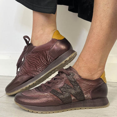  metallic bronze-brown leather sneakers with a textured chevron pattern, dark glittery star logo, and mustard yellow heel tabs, shown on a person's feet.
