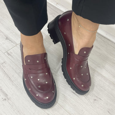 Burgundy leather shoes with studs worn indoors on a light wooden floor.