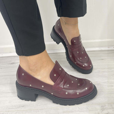 Burgundy loafers with stud details worn by a person on a light wooden floor.