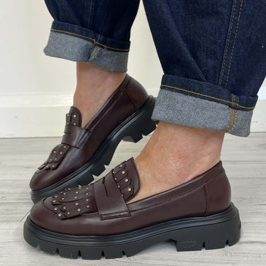 A person wearing dark brown leather loafers with studded straps and chunky black lug soles, paired with dark blue cuffed jeans, standing on a light wood floor.