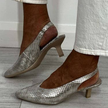 Silver high-heeled shoes worn with white pants on a wooden floor.