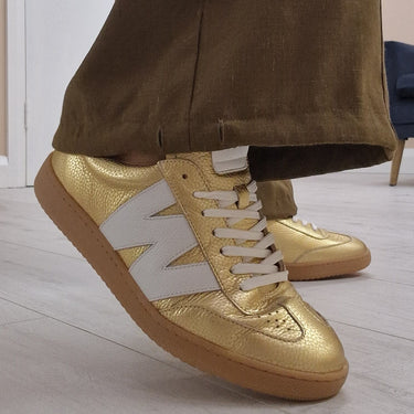 Gold sneakers with white laces worn with brown pants on a light wooden floor.
