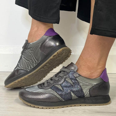 A pair of low-top sneakers shown on a person's feet. The shoes have a metallic dark gray and blue-purple textured upper with a purple heel tab and a tan outsole.