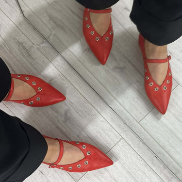 Red pointed-toe shoes with studded details worn by a person on a light wooden floor.
