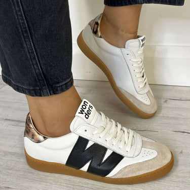 White sneakers with black and gold accents worn with dark jeans on a light wooden floor.