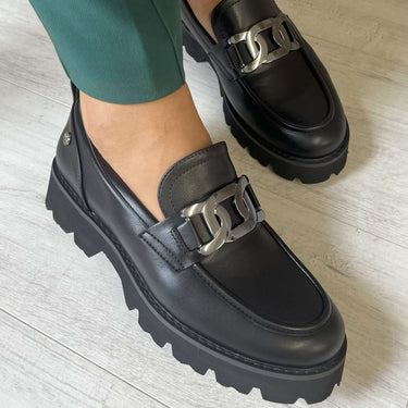 Black loafers with silver buckles worn with green pants on a light wooden floor.