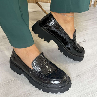 Black crocodile-patterned loafers worn with green pants on a light wooden floor.