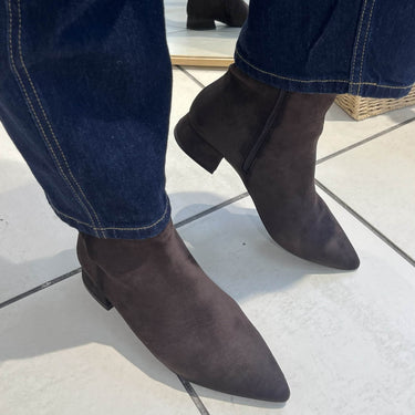 A person wearing dark brown pointed-toe ankle boots with low block heels, paired with dark blue jeans, standing on a light tiled floor.