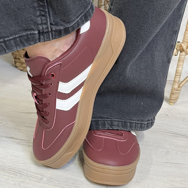 Maroon sneakers with white stripes and brown soles worn with blue jeans on a light wooden floor.