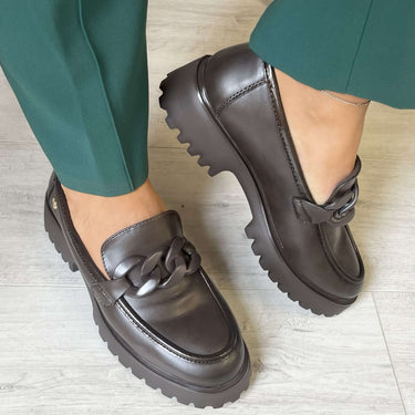 Brown loafers worn with green pants on a light wooden floor.