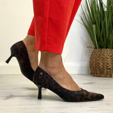 Person wearing red pants and black high-heeled shoes on a light wooden floor with a plant in the background.