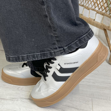 silver sneakers with brown soles worn with dark jeans on a light wooden floor.