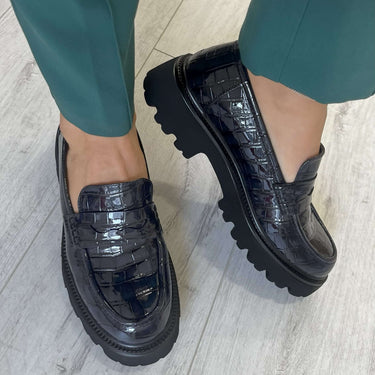 navy loafers worn with green pants on a light wooden floor.