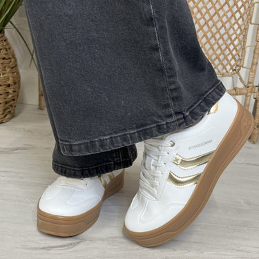 White sneakers with brown soles worn with dark jeans on a light wooden floor.
