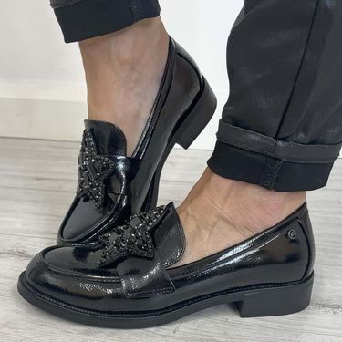Black patent leather shoes with embellishments worn by a person on a light wooden floor.